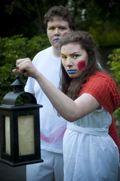 Julie is holding a lantern in front of her. She wears a white dress with  a red scarf draped around her shoulders. She has bright yellow, blue and red face paint. Behind her, Francis stands dressed in white with the same colourful face paint. 