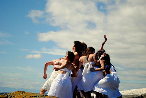 A cluster of performers dressed in white cloth are pressing against one another. They are on a rock in the ocean and there is a cloudy sky behind them. 