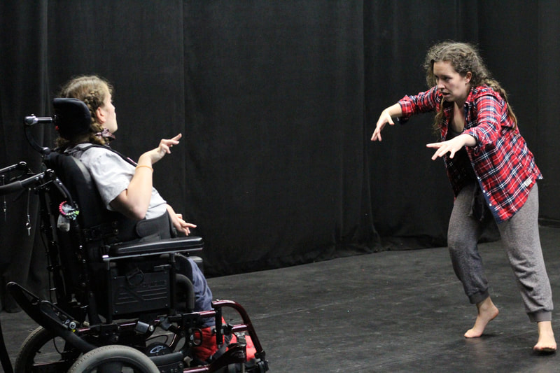 Two dancers face each other with their arms raised. One uses a manual wheelchair and the other is standing. 